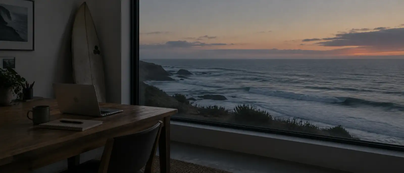 Desk with a laptop and a surfboard by a large window overlooking the Atlantic at dusk